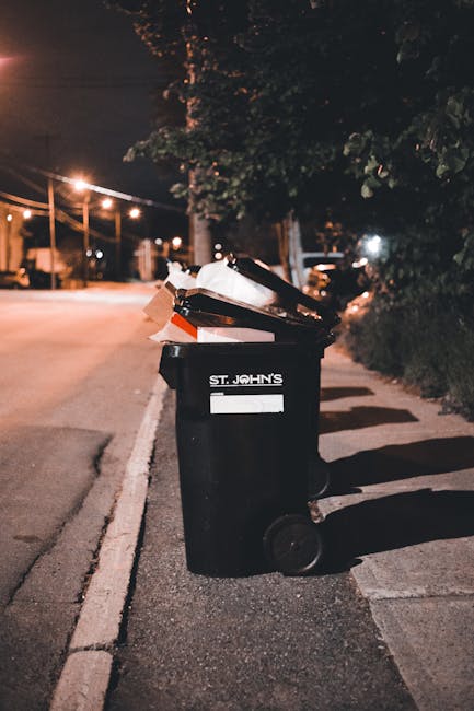 A black wheeled rubbish bin labeled 'St. John's' positioned on a paved sidewalk next to a curb along a quiet street at night. The lid of the bin is open, revealing a mixture of discarded cardboard boxes, paper, and other domestic waste. The street is illuminated by warm streetlights, casting soft light and shadows onto the pavement and the bin. In the background, there are trees with dark foliage, and faint outlines of parked cars and buildings are visible in the distance, indicating an urban or suburban environment. The scene suggests an independent collection or private waste disposal scenario outside a residential property, with the bin ready for rubbish removal services. The overall atmosphere is calm, with a focus on routine waste management practices, reflecting the type of visible waste accessible for scheduled rubbish removal by companies such as Rubbish Removal Barnes.