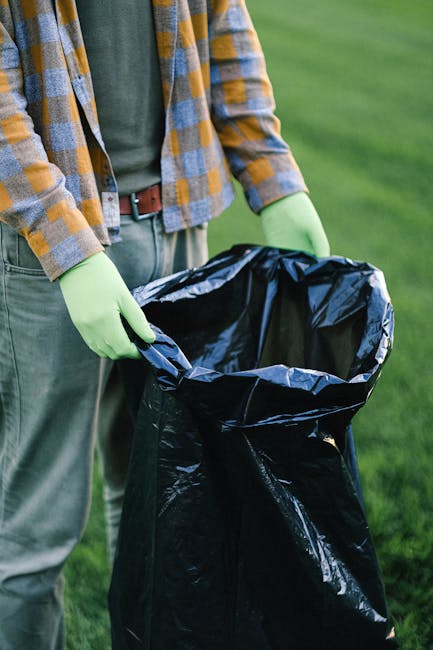 A collection of assorted rubbish and waste materials are piled on the ground in an outdoor setting. The debris includes black plastic garbage bags, a yellow plastic container, a used car tire, and various loose items such as paper, plastic, and packaging. The waste is situated on a gravel surface in front of a low stone wall made of uneven, roughly cut stones. In the background, there is a metal utility pole with wires extending across the sky, which is partly cloudy with patches of blue visible. Beyond the wall, a fenced area contains a large, semi-collapsed dirt-covered structure or tent with a white metal frame, and some greenery including small trees or bushes. The scene appears to be an informal or unmanaged waste accumulation site, highlighting the importance of scheduled rubbish removal services like those offered by Rubbish Removal Barnes, which specialises in private disposal and alternative waste handling options outside municipal collection.