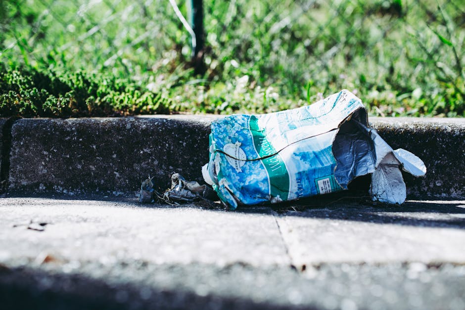A crumpled plastic bag and trash lying on an outdoor concrete pavement near the curb of a street, with surrounding green grass and foliage in the background. The plastic bag features a blue and white design with some text and images, indicating it is a used shopping or grocery carrier. The bag appears to be damaged, torn, and partially flattened on the ground, with small pieces of debris nearby. The scene is illuminated by natural daylight, casting shadows on the pavement. This image highlights the importance of proper rubbish disposal and the role of rubbish removal services, such as those provided by Rubbish Removal Barnes, in maintaining cleanliness and environmental care in urban and residential areas, especially during private waste collection or on-site clearance activities.