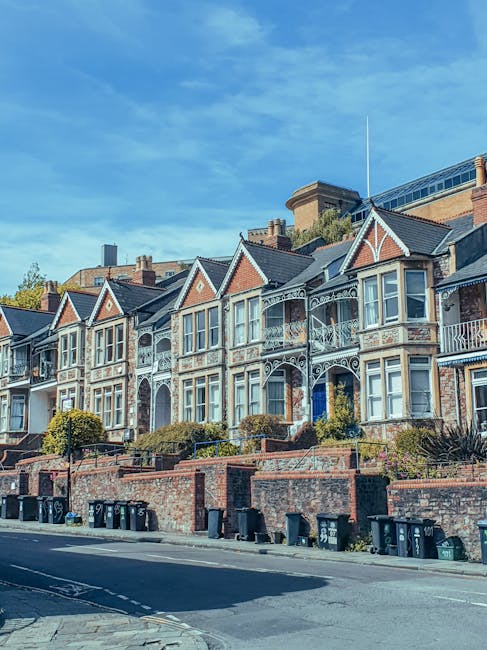 A row of historic Victorian-style terraced houses situated along a sloped street in Barnes, London. The houses feature ornate white ironwork balconies, large bay windows, and decorative brickwork with varied gable rooflines. The buildings are set behind a low brick retaining wall with small landscaped front gardens, some with trimmed shrubs and plants. The pavement in the foreground is lined with multiple black wheelie bins arranged near the curb, likely for waste collection. The street appears quiet, with no visible vehicles or pedestrians, under a clear blue sky with some wispy clouds. The scene may relate to private waste handling or alternative rubbish disposal options, with Rubbish Removal Barnes occasionally involved in on-site clearance services for such residential properties.
