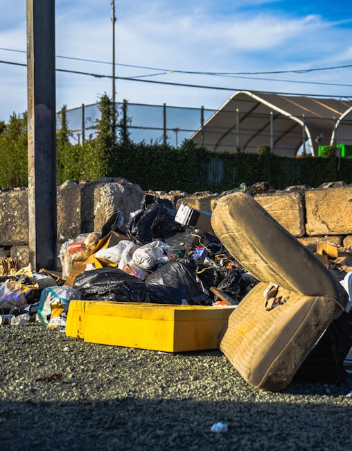 A pile of mixed household rubbish and waste materials is seen scattered on the ground next to a low stone wall in an outdoor setting. The waste includes black plastic garbage bags, a yellow plastic storage container, and a discarded car tire leaning against the pile. The surface appears to be gravel or asphalt, with some loose debris visible. In the background, there is greenery, such as bushes and trees, behind a metal fence and a tennis court or sports facility with a curved metal roof structure. A wooden utility pole with overhead wires extends vertically in the foreground, and the scene is illuminated by daylight under a mostly clear blue sky. The image showcases an example of unmanaged waste possibly requiring private rubbish removal services, highlighting the importance of proper disposal of household or garden refuse in the Barnes area.