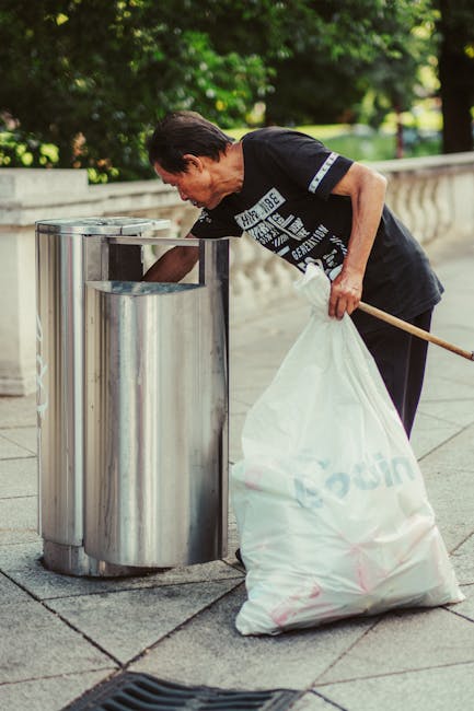 A middle-aged man with dark hair, wearing a black T-shirt with white text and dark pants, is seen disposing of rubbish into a public metal trash bin on a paved sidewalk. The trash bin is cylindrical, made of stainless steel, with a rounded top and an opening at the front where the man is placing a white plastic bag filled with waste. He is slightly bent forward, holding the plastic bag in his right hand and steadying a wooden stick in his left hand, which is resting on his waist. The background features lush green trees and a stone balustrade, suggesting a park or similar outdoor setting. The scene is well-lit, indicating daytime, and the environment appears clean and well-maintained. The image visually supports the context of independent rubbish disposal and litter management, relevant to private waste handling and rubbish removal services, as provided by Rubbish Removal Barnes.
